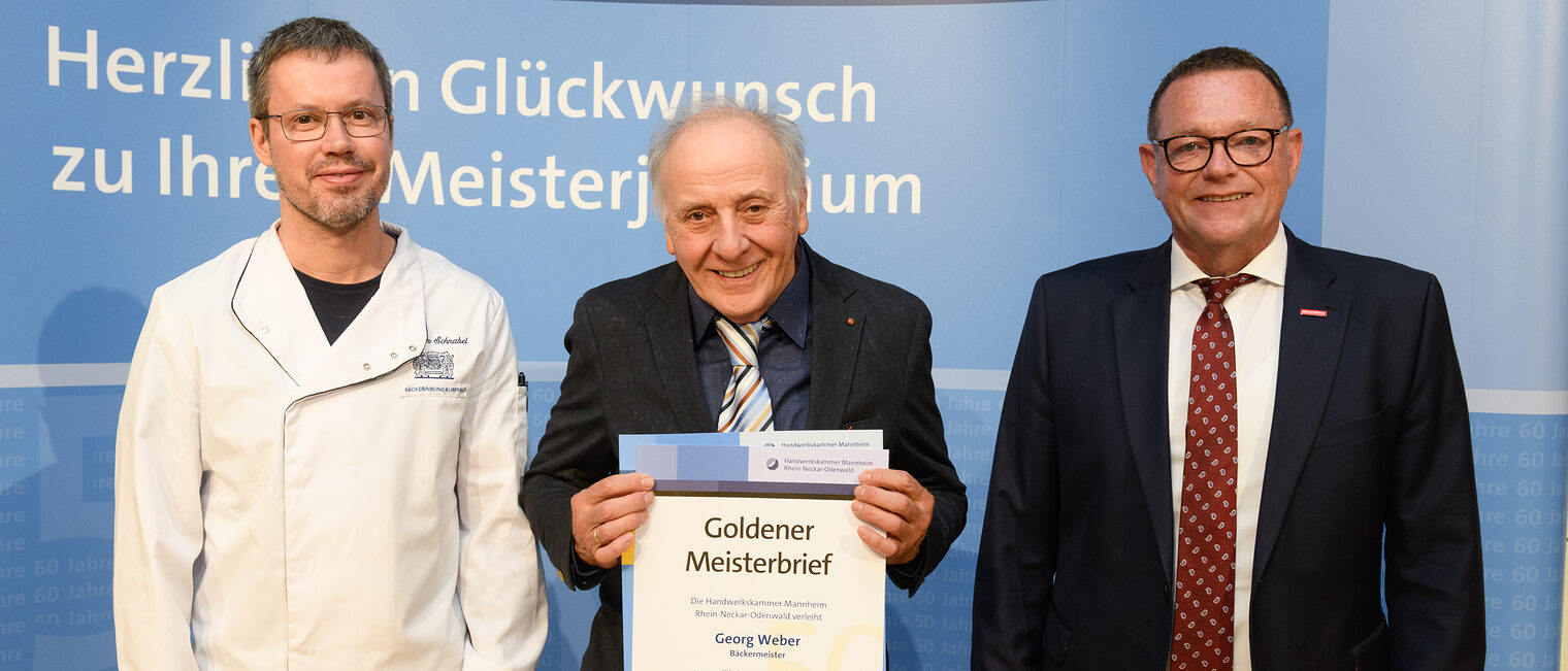 Drei Personen posieren für ein Foto, in der Mitte wird die Ehrenurkunde gehalten