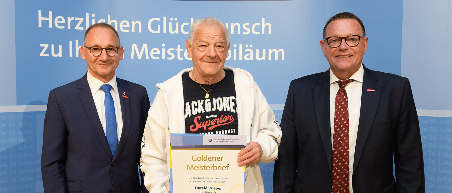 Drei Personen posieren für ein Foto, in der Mitte wird die Ehrenurkunde gehalten