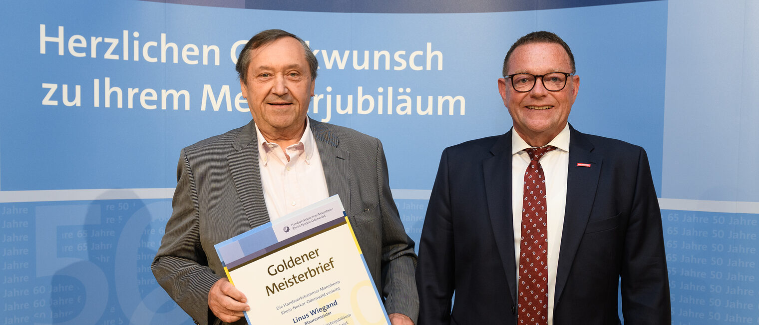 Zwei Personen posieren für ein Foto, in der Mitte wird die Ehrenurkunde gehalten