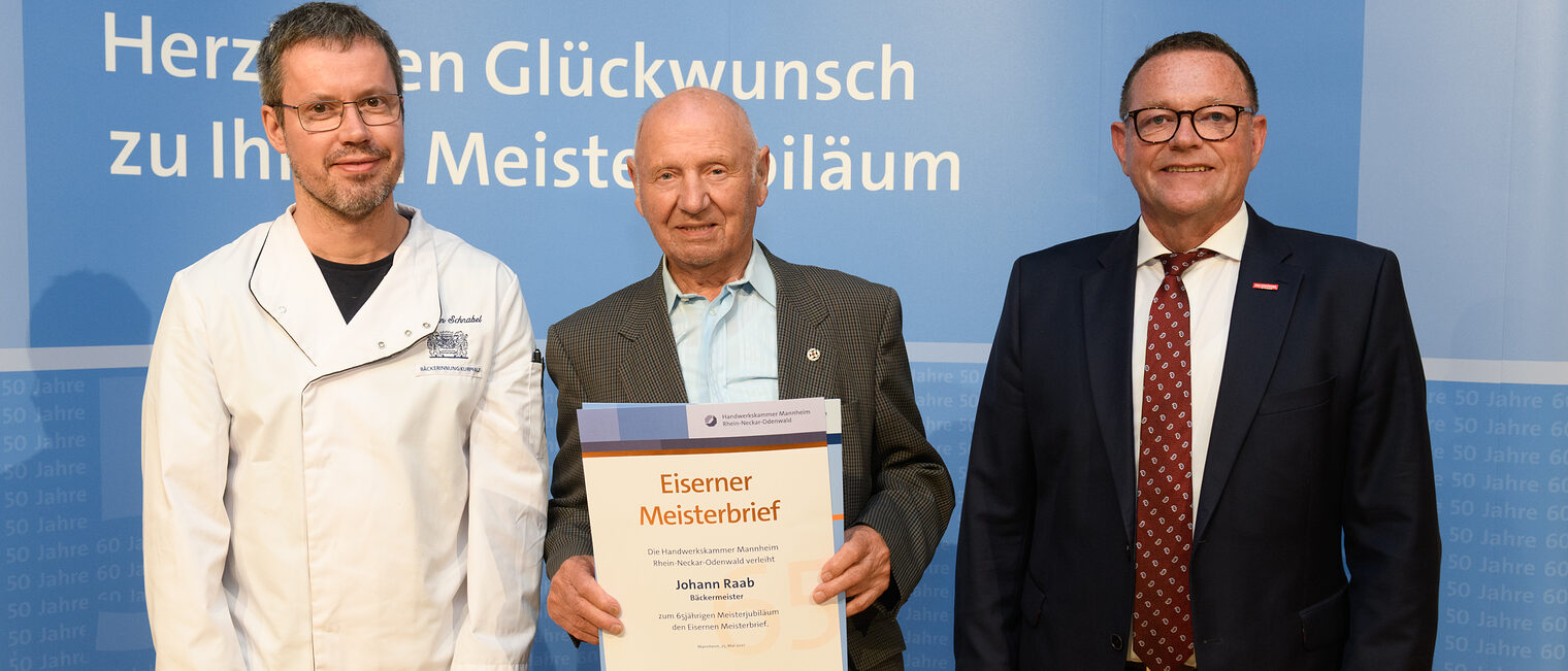 Drei Personen posieren für ein Foto, in der Mitte wird die Ehrenurkunde gehalten