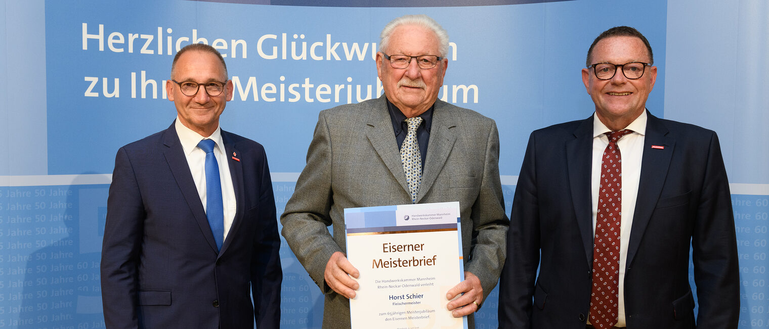 Drei Personen posieren für ein Foto, in der Mitte wird die Ehrenurkunde gehalten