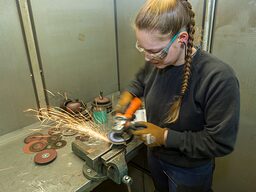 Eine Frau mit der Flex am bearbeiten eines Metallstücks im Schraubstock