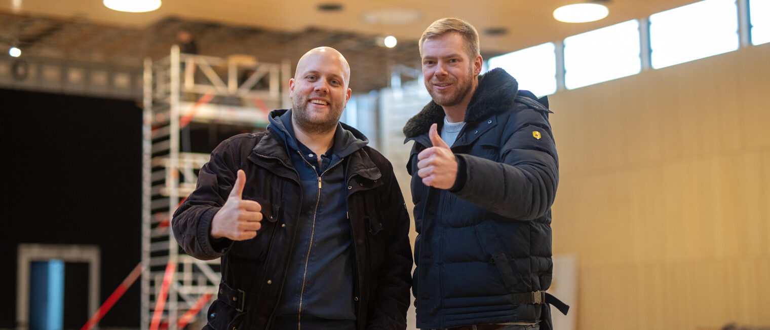 Zwei Handwerker in Winterjacken stehen lächelnd nebeneinander in einer Halle und zeigen beide den Daumen hoch, im Hintergrund ein Baugerüst und große Fenster.