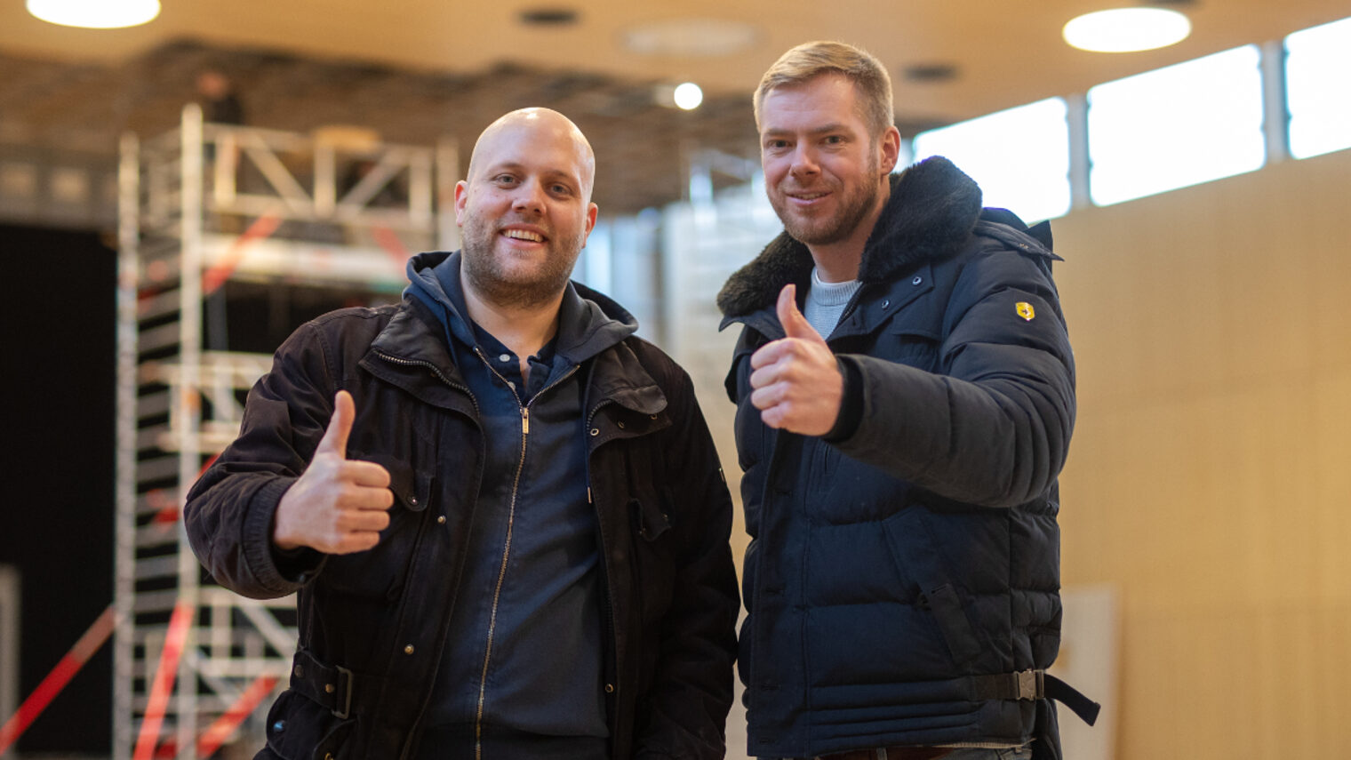 Zwei Handwerker in Winterjacken stehen lächelnd nebeneinander in einer Halle und zeigen beide den Daumen hoch, im Hintergrund ein Baugerüst und große Fenster.