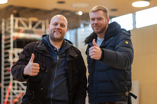 Zwei Handwerker in Winterjacken stehen lächelnd nebeneinander in einer Halle und zeigen beide den Daumen hoch, im Hintergrund ein Baugerüst und große Fenster.