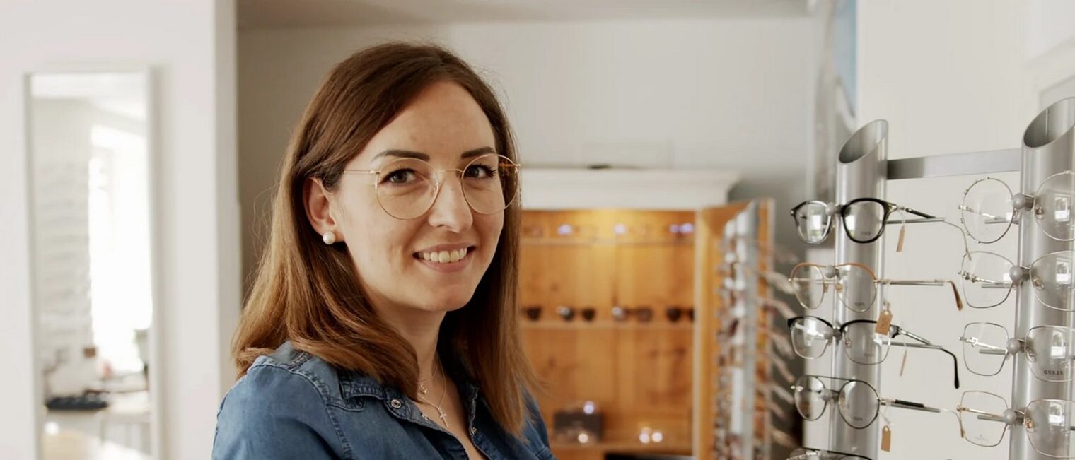 Eine Frau mit einer Brille schaut in die Kamera und steht vor einer Auswahl an Brillen