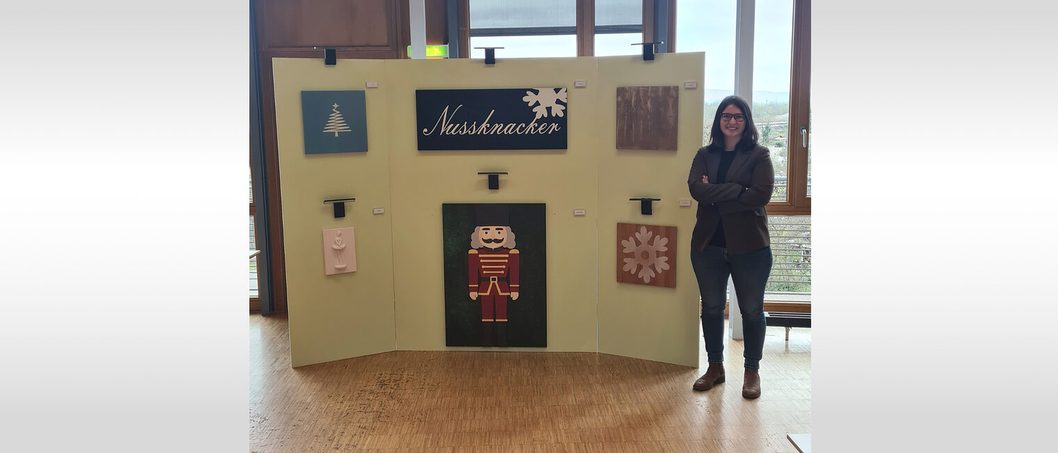 Frau steht neben einer Ausstellung Ihrer Malerarbeit mit Weihnachtsbaum, Nussknacker und weiteren Holzkunstwerken.