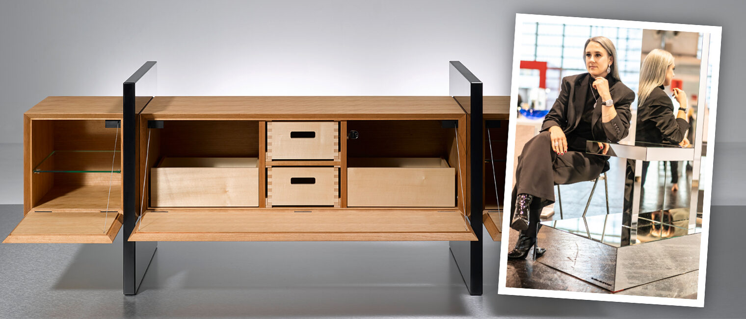 Schreinerin Nicole Doth sitzt neben ihrer Spiegelskulptur; links das geöffnete Holz-Sideboard „3hoch2“ mit schwarzen Seitenwangen.