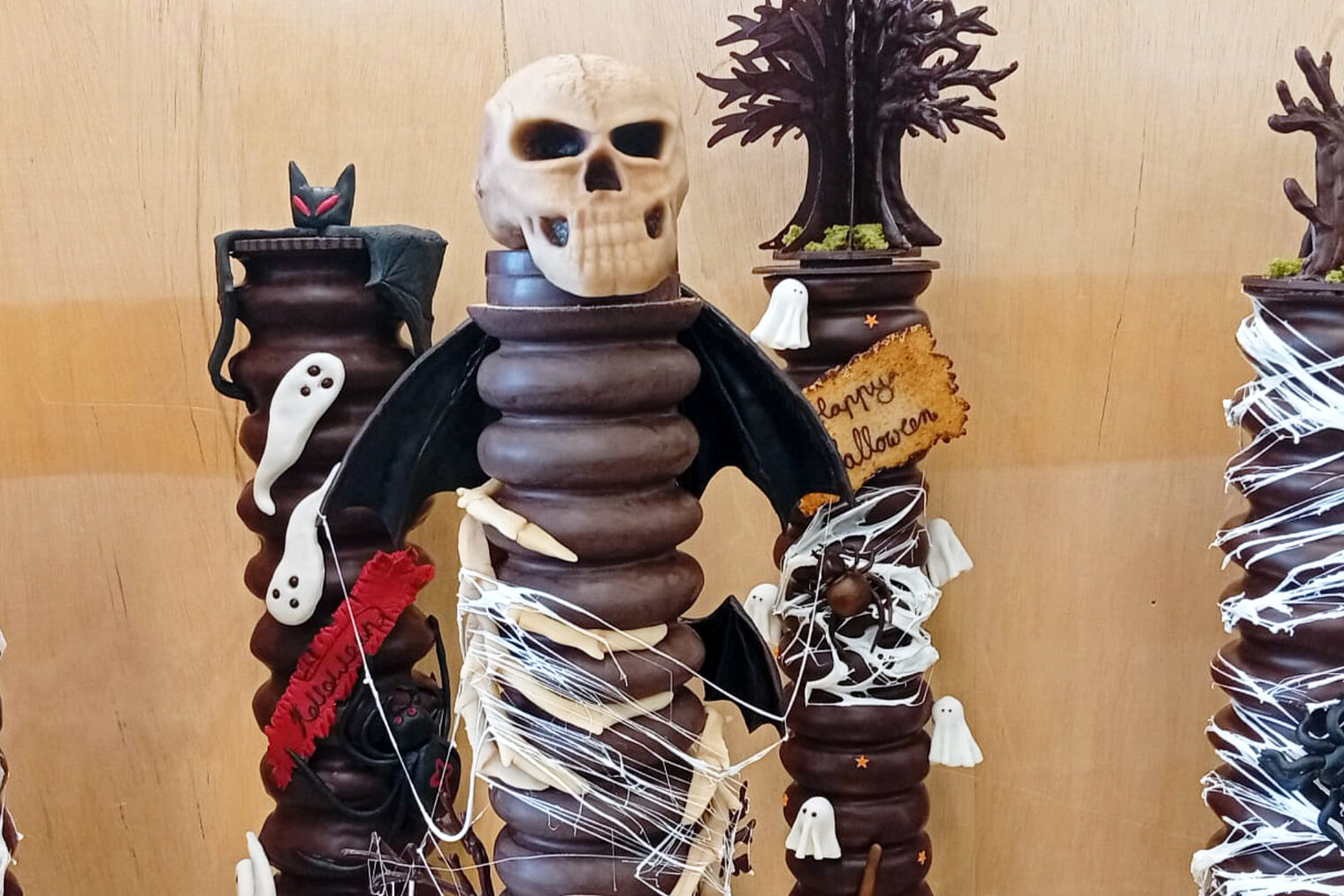 Präsentation von sechs mit dunkler Schokolade überzogene Baumkuchen vor einer Holzwand, die Kuchen tragen Gruselmotive zu Halloween wie Fledermäuse, Spinnen, Totenschädel, Kürbise und andere Halloween-Motive