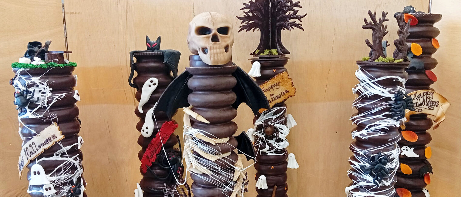 Präsentation von sechs mit dunkler Schokolade überzogene Baumkuchen vor einer Holzwand, die Kuchen tragen Gruselmotive zu Halloween wie Fledermäuse, Spinnen, Totenschädel, Kürbise und andere Halloween-Motive